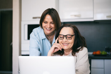 Adult daughter and her mature mother looking at the laptop screen and laughing while making online...