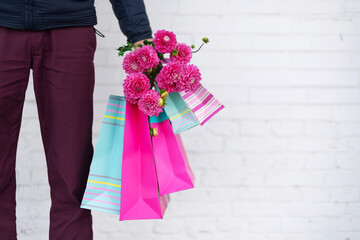 Stylish man holding flowers and pink, blue shopping bags on brick background. Woman's day. Valentine's day. Ready for birthday party or romantic date. Copy space. Bunch of paper gift bags