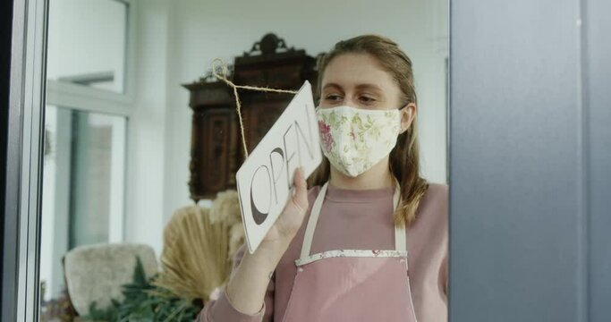 A young woman who owns a small business dressed in a mask turns over a sign openly and opens her flower shop during quarantine and the virus