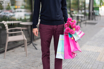 Romantic man with pink flowers and shopping bags on brick background. Woman's day. Valentine's day. Ready for birthday party or romantic date. Copy space. Bunch of paper gift bags