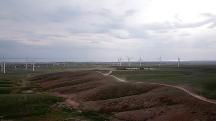 Windmills stand in the middle of green fields. Clean energy is generated. Cloudy weather, dark clouds. Green grass grows. High hills in the distance. Top view from drone. Industrial farm of Kazakhstan