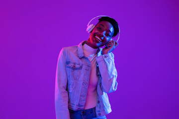 Happy young African American woman wearing headphones listening to music on  purple neon light background