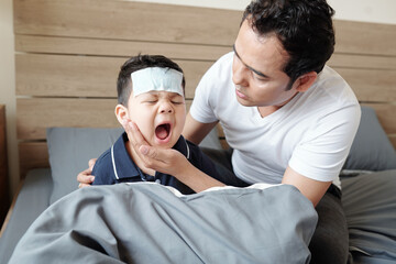 Fototapeta premium Worried father sitting in bed next to his kid and comforting little son crying from fever