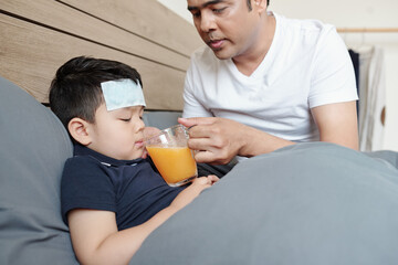 Caring father giving glass of fresh orange juice to his sick little kid lying in bed with cooling...