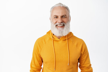 Close up of happy handsome old man with beard, smiling and looking cheerful at camera, standing in...