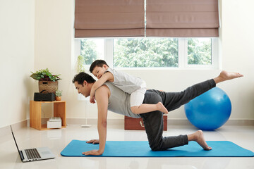 Laughing cute little boy interfering his dad following tutorial on laptop screen when practicing...