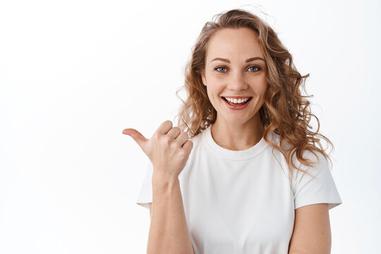 Image of stylish blond woman points left, shows promotional text aside, smiles and looks satisfied at camera, white background