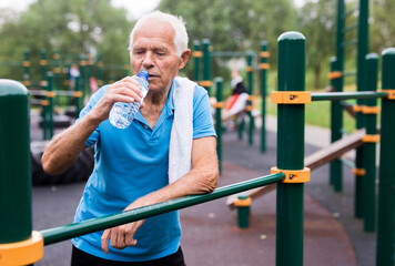 Obraz premium old peppy pensioner man resting and drinking water on a sports equipped playground