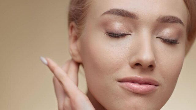 Female beauty. Beautiful young blonde lady touching her face and smiling to camera, beige studio background, empty space