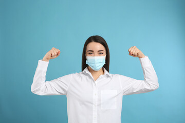 Woman with protective mask showing muscles on light blue background. Strong immunity concept