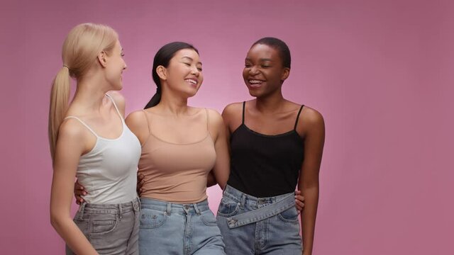 Great shopping promo. Three diverse women looking aside and laughing at empty space, smiling to camera, pink background