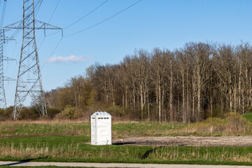 Isolated white portable toilet at the end of a parking lot and beside a field and hydro electric towers. Forest in the background. 
