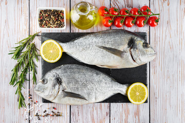 Fresh fish dorado on black slate cutting board and white wooden background with ingredients for cooking