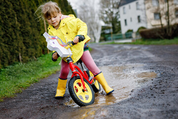 Little toddler girl wearing yellow rain gum boots, running with balance bike during sleet. Happy child driving, biking with bicycle into puddle, splashing water, outdoor activity. Happiness, childhood