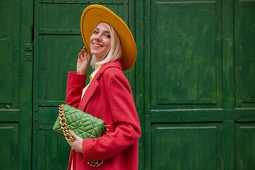Fashionable happy smiling woman wearing yellow hat, cpral color coat, holding quilted faux leather...