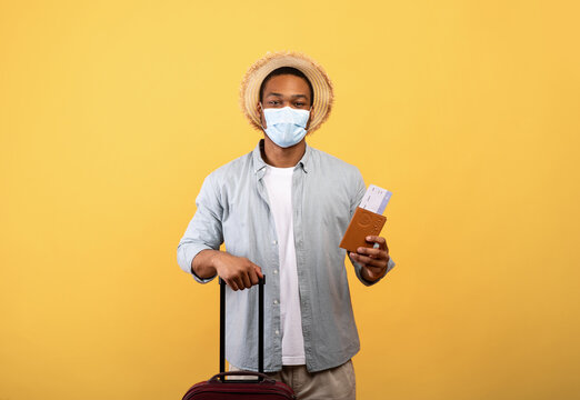 African American Man In Medical Mask Holding Passport, Airplane Tickets And Luggage On Orange Background