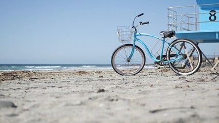 Naklejka na ściany i meble Blue bicycle, cruiser bike by ocean beach, pacific coast, Oceanside California USA. Summertime vacations, sea shore. Vintage cycle on sand near lifeguard tower or watchtower hut. Sky and water waves.