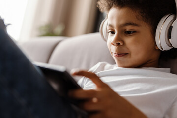 Black curly boy in headphones using tablet computer while sitting on sofa