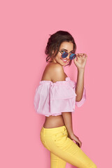 Beautiful girl in yellow jeans and pink shirt, with hands up wearing sunglasses posing, dancing, smiling on pink background in studio.