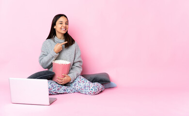 Young mixed race woman eating popcorn while watching a movie on the laptop pointing finger to the side