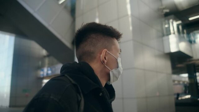 Young Asian man with short hair in protective medical mask walks down the street of modern city. Exit from airport or train station. Sunny day and walk outside using a face mask. Black denim jacket