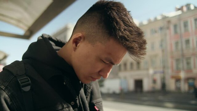 young Asian man stands at the bus stop and looks at his cell phone. Tracking city transport using a mobile device. City application. Modern infrastructure. Being late for public transport