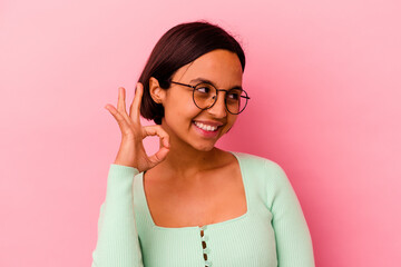 Young mixed race woman isolated on pink background winks an eye and holds an okay gesture with hand.