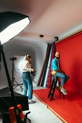 Fototapeta na wymiar Woman photographer making portraits for handsome bearded man in photo studio on red background, backstage photo. Vertical