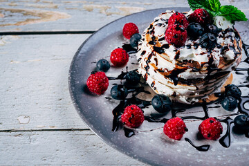 plate of stack of pancakes with cream and chocolate syrup with blueberries, strawberries and raspberries on rustic wooden table.
