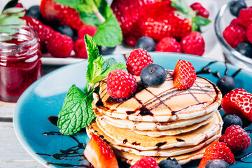delicious and colorful plate of pancakes with fresh fruits and mint leaves dipped in chocolate syrup. small jar of strawberry jam in the background.