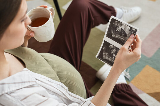 Young pregnant woman enjoying the cup of tea and looking at ultrasound of her baby while sitting on the sofa