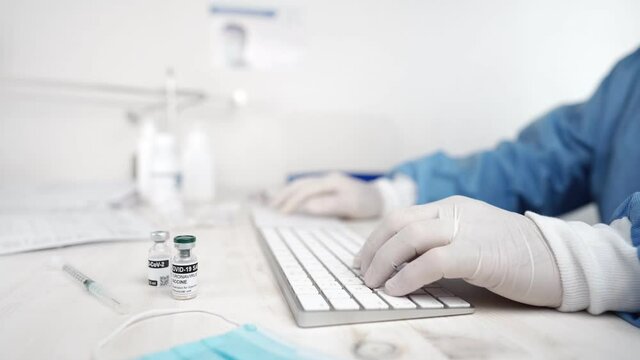 Close up of a doctor or healthcare worker in personal protective kit typing on pc keyboards with syringe, medical stuff and ampoules of Covid-19 Coronavirus vaccine in foreground in selective focus