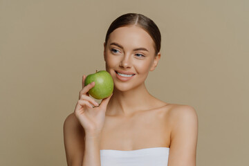 Thoughtful charming European woman holds apple near face smiles gently has white perfect teeth healthy clean skin wrapped in shower towel stands with bare shoulders against brown background.