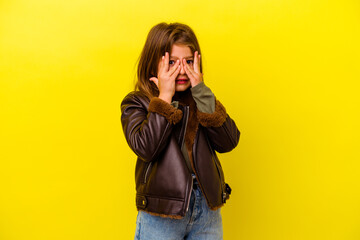 Little caucasian girl isolated on yellow background blink through fingers frightened and nervous.