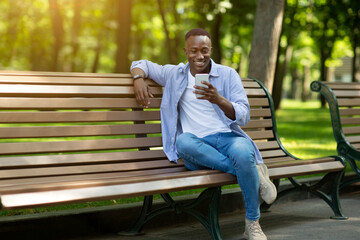 Smiling black guy sitting on bench at park and using his smartphone, copy space