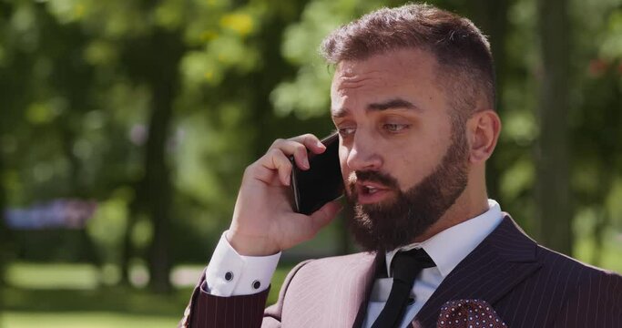 Emotional bearded businessman talking with problems via mobile phone, solving business problems during break at park