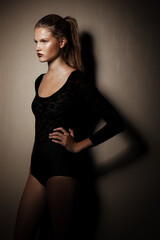 Fototapeta premium Female model in black bodysuit with tight hair in the ponytail, posing in the studio, looking in profile, dark background.