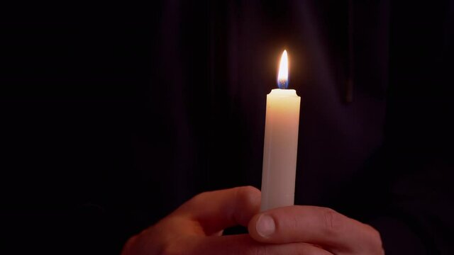 Male Holds in Hands a Lighted Wax Candle in a Dark Room. Bright flame, fire, ignition. Supplication. White paraffinic candle with yellow fire. Concept of memory, ritual, celebration. 4K.