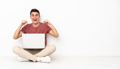 Teenager man sitting on the flor with his laptop celebrating a victory in winner position
