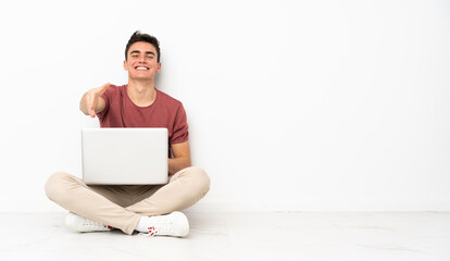 Teenager man sitting on the flor with his laptop shaking hands for closing a good deal