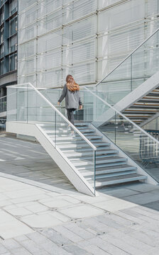 Mujer pelirroja vestida con pantalones negros y chaqueta gris subiendo unas escaleras de acero blancas con barandillas de vidrio azulado, frente a un edificio blanco de fachada met&aacute;lica un soleado d&iacute;a