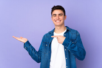 Teenager caucasian  handsome man isolated on purple background holding copyspace imaginary on the...