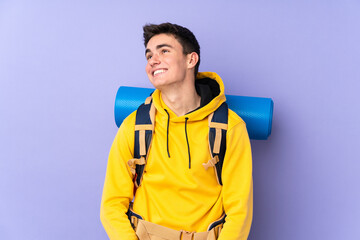 Teenager caucasian mountaineer man with a big backpack isolated on purple background laughing and looking up