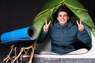 Teenager caucasian man inside a camping green tent isolated on black background showing victory sign with both hands