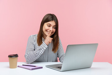 Please, keep secret. Positive woman in striped shirt shushing with silence gesture, talking on video call, having conference via laptop at home office. Indoor studio shot isolated on pink background