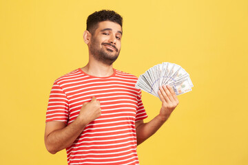 Egoistic man with beard in striped t-shirt pointing finger at himself holding fan of hundred dollar...