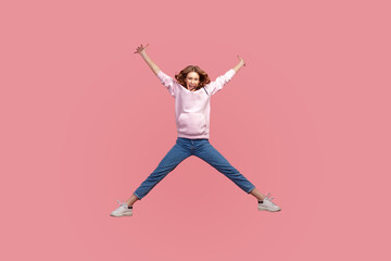 Full length portrait of glad young female in hoodie and jeans jumping high in air, flying and shouting with amazed happy expression, celebrating success. Indoor studio shot isolated on pink background
