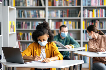 Students during pandemic with medical masks. Concentrated african american girl wears protective medical mask sit at table in library, preparing for exam, takes notes, classmates sit at background