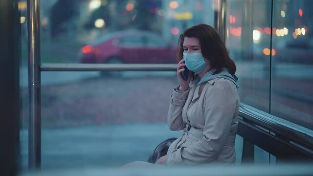 Woman in mask sitting at bus stop and talking on phone, shot through glass, cars driving on blurred background. Handheld elegant female having conversation while waiting for transport