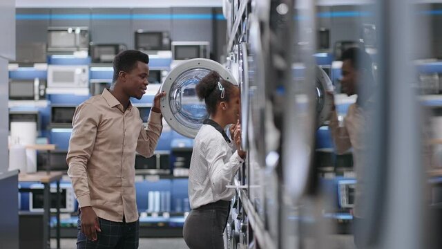 cheerful afro-american couple is choosing washing machine in hardware shop, man and woman are viewing new models inside